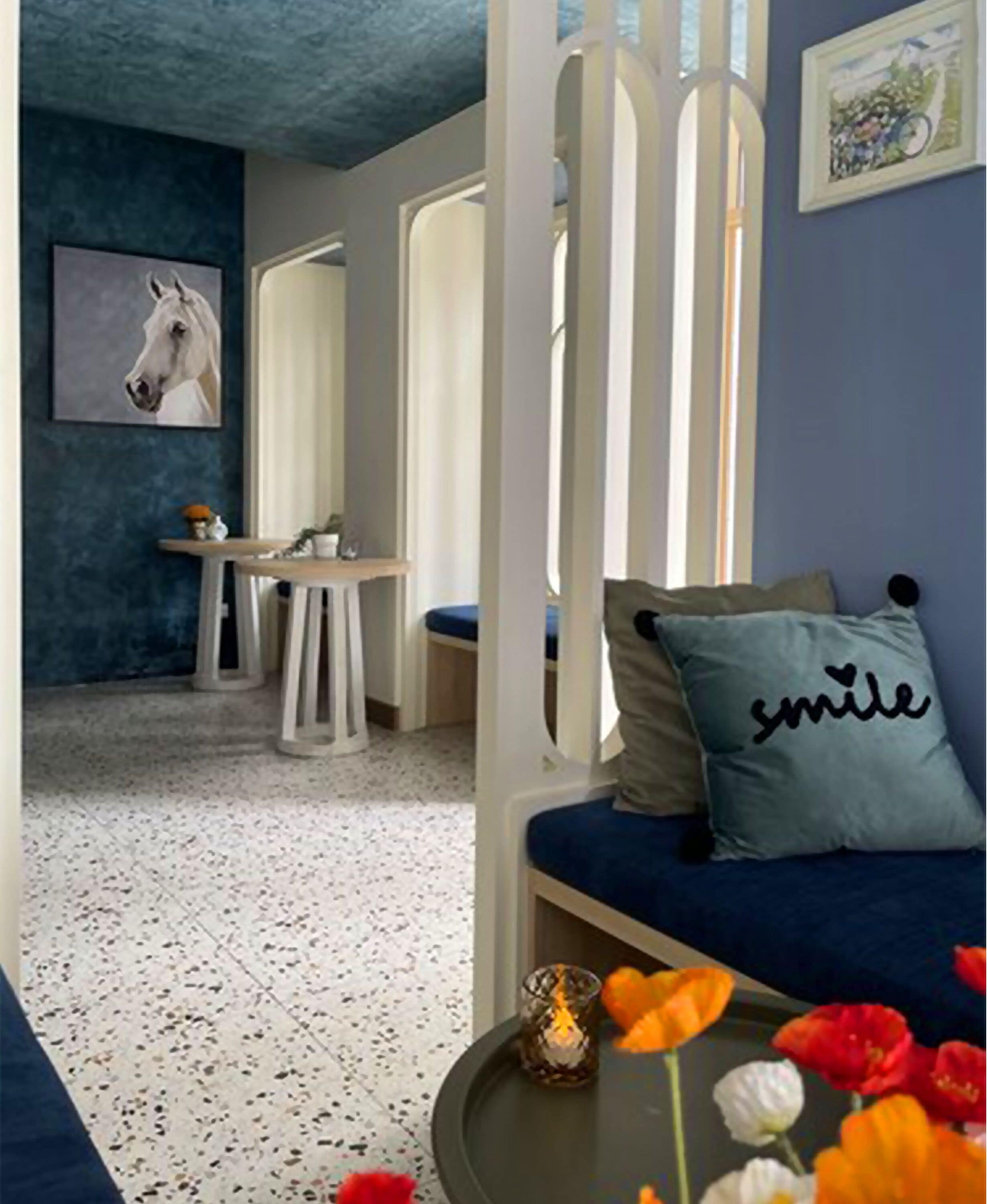 Et hyggeligt rum med blå vægge, hvidt loft og en blå sofa med en pude, der siger "smile". Der er et lille bord med et stearinlys og blomster, samt et maleri af en hest på væggen.