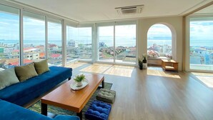 Un salón espacioso con un ventanal que ofrece vistas a la ciudad, un sofá azul, una mesa de centro de madera y una pequeña zona de estar con un banco.