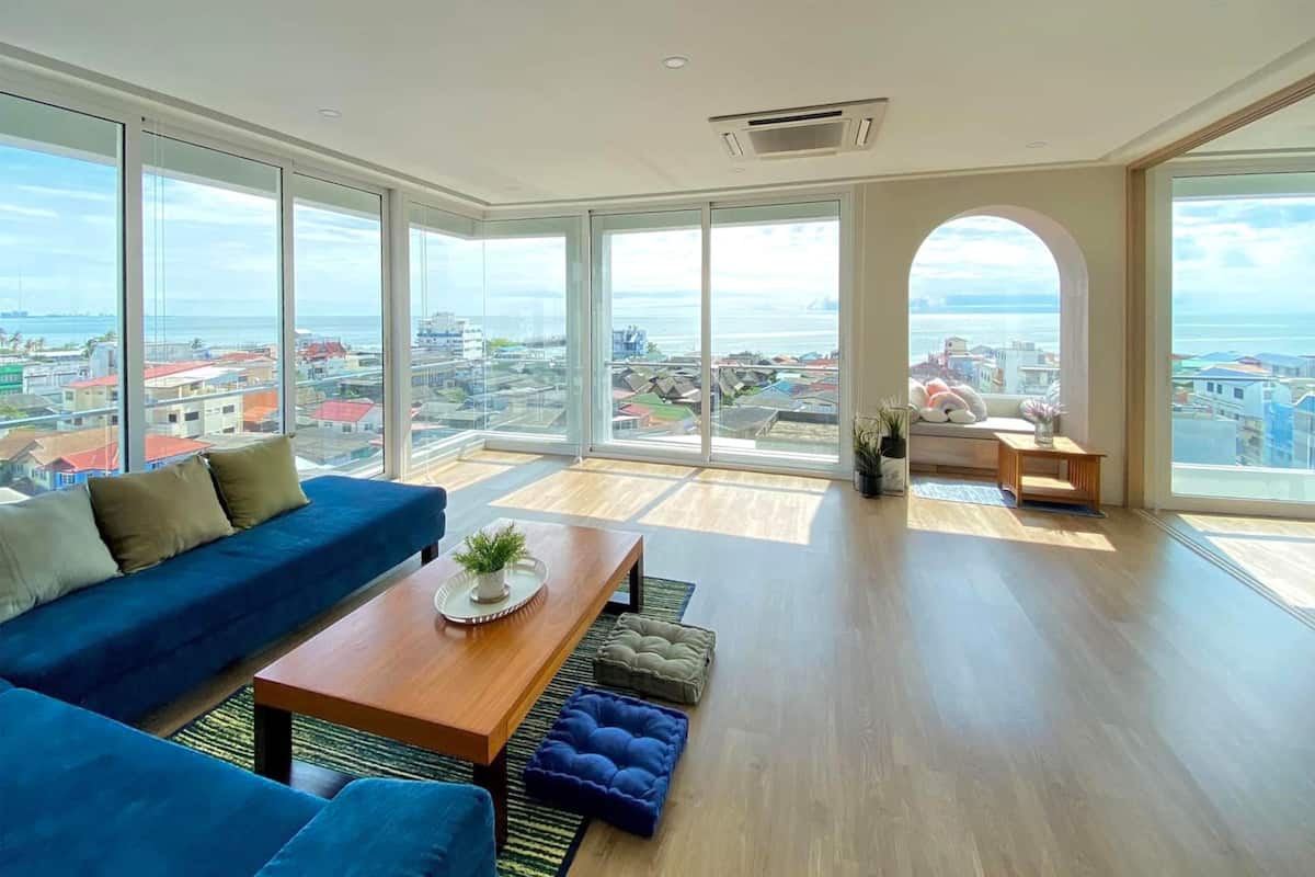 A spacious living room with a large window offering a city view, a blue sofa, a wooden coffee table, and a small seating area with a bench.