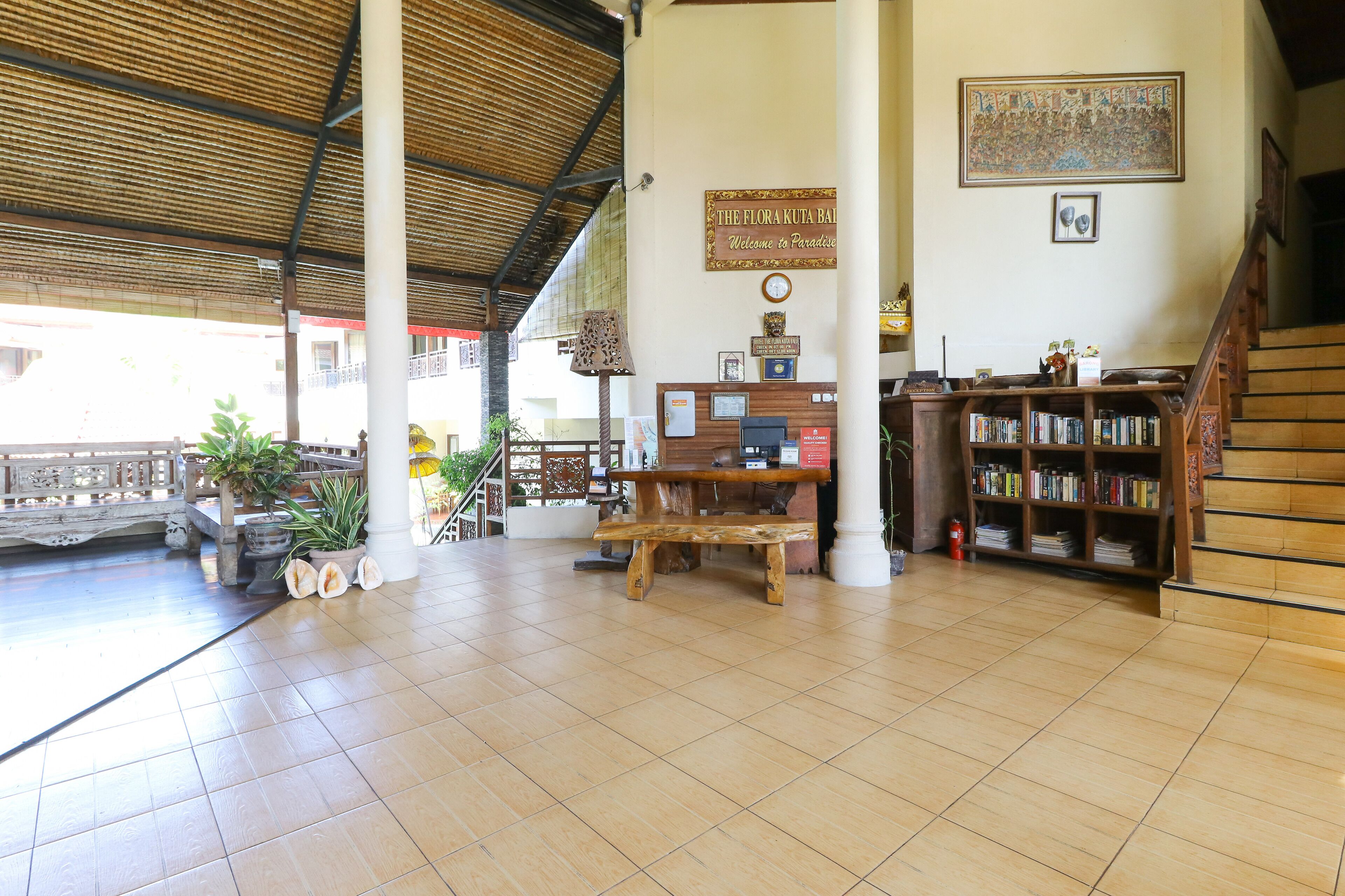 Uma área de recepção com um banco de madeira, uma estante, uma placa com a inscrição "The Flora Kuta Bali Bem-vindo ao Paraíso" e uma escada.