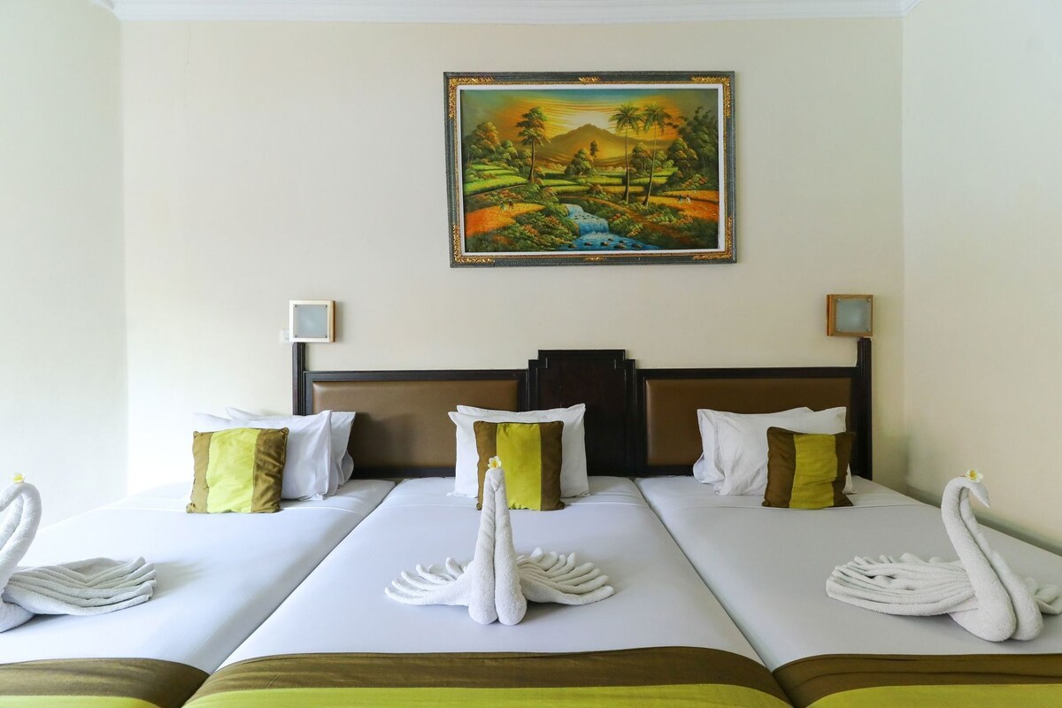 A hotel room with two beds, each with a swan towel and pillows, a painting of a landscape on the wall, and a small table lamp.