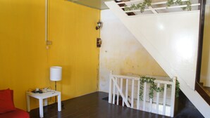 Una habitación con una pared amarilla, una escalera blanca, un sofá rojo, una pequeña mesa blanca con una lámpara y una barandilla blanca adornada con plantas.