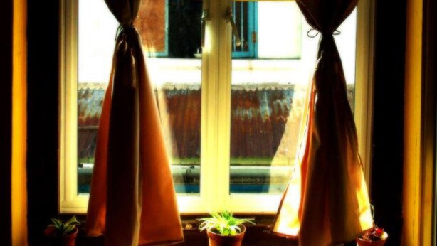 Una ventana con cortinas atadas en la parte superior, una pequeña planta en maceta en el alféizar y un letrero con caracteres chinos.