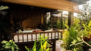 A covered outdoor seating area with a wooden bench, surrounded by various plants and a bamboo roof structure. - Noordin Street House (George Town)