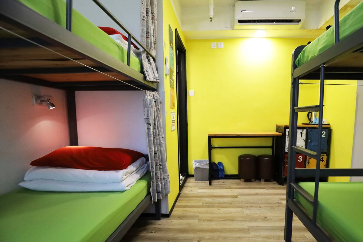 A dormitory room with bunk beds, a small table, and a trash bin.