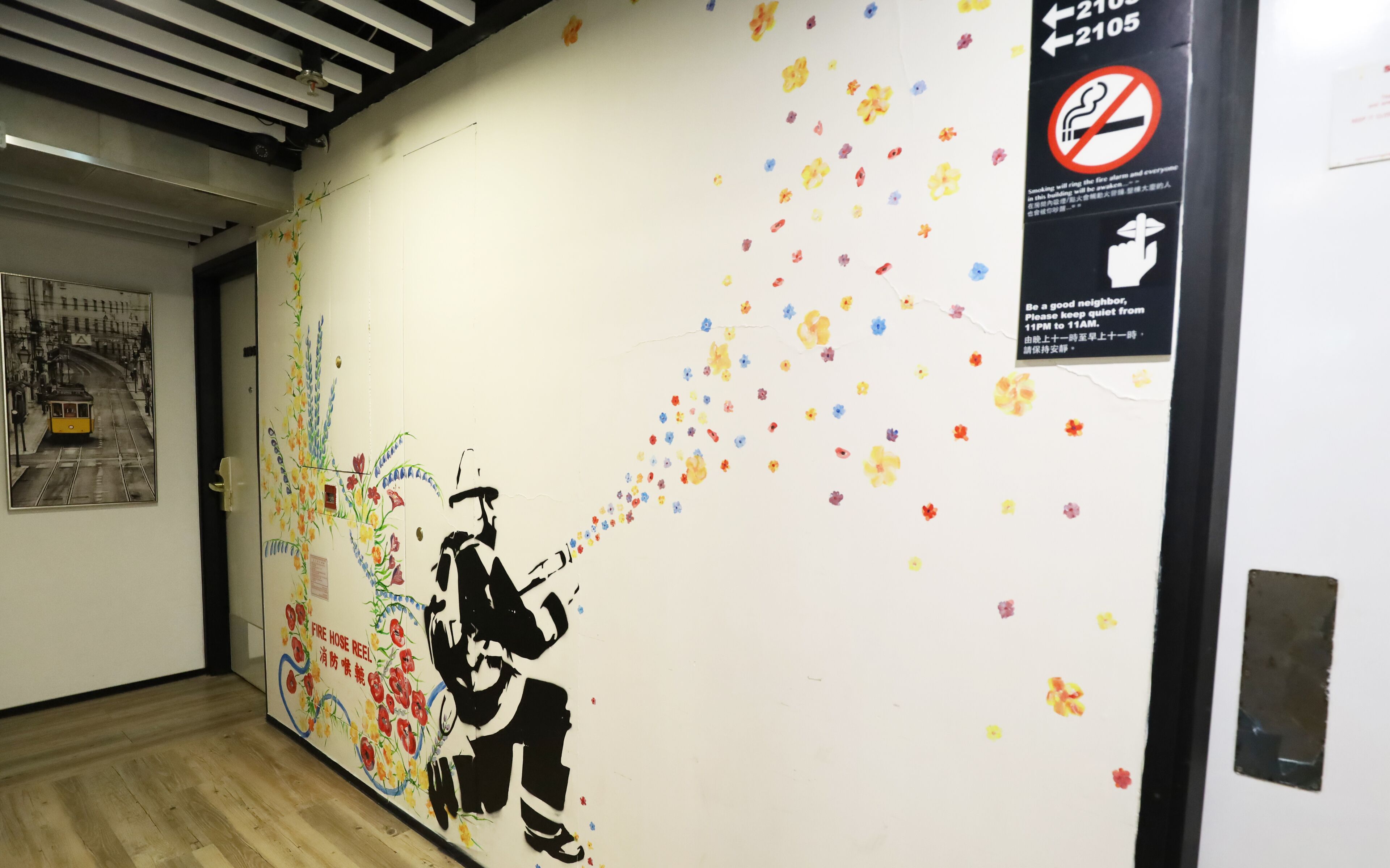 Un pasillo con un mural de una persona manejando una manguera contra incendios, un letrero de prohibido fumar y un cartel pidiendo ser un buen vecino.