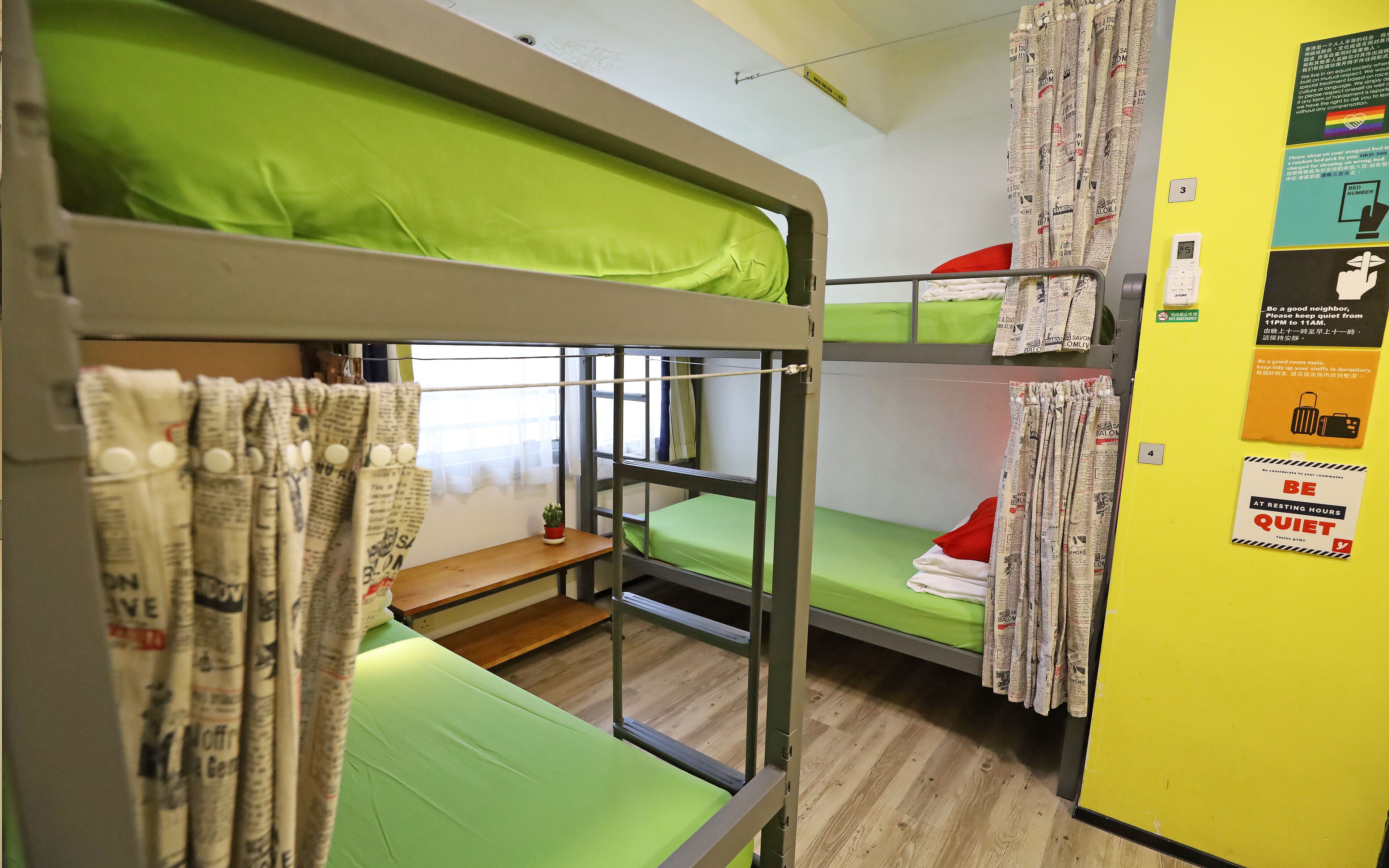 A bunk bed room with green bedding, a small table, and a red pillow. There are curtains made of newspaper on the windows.