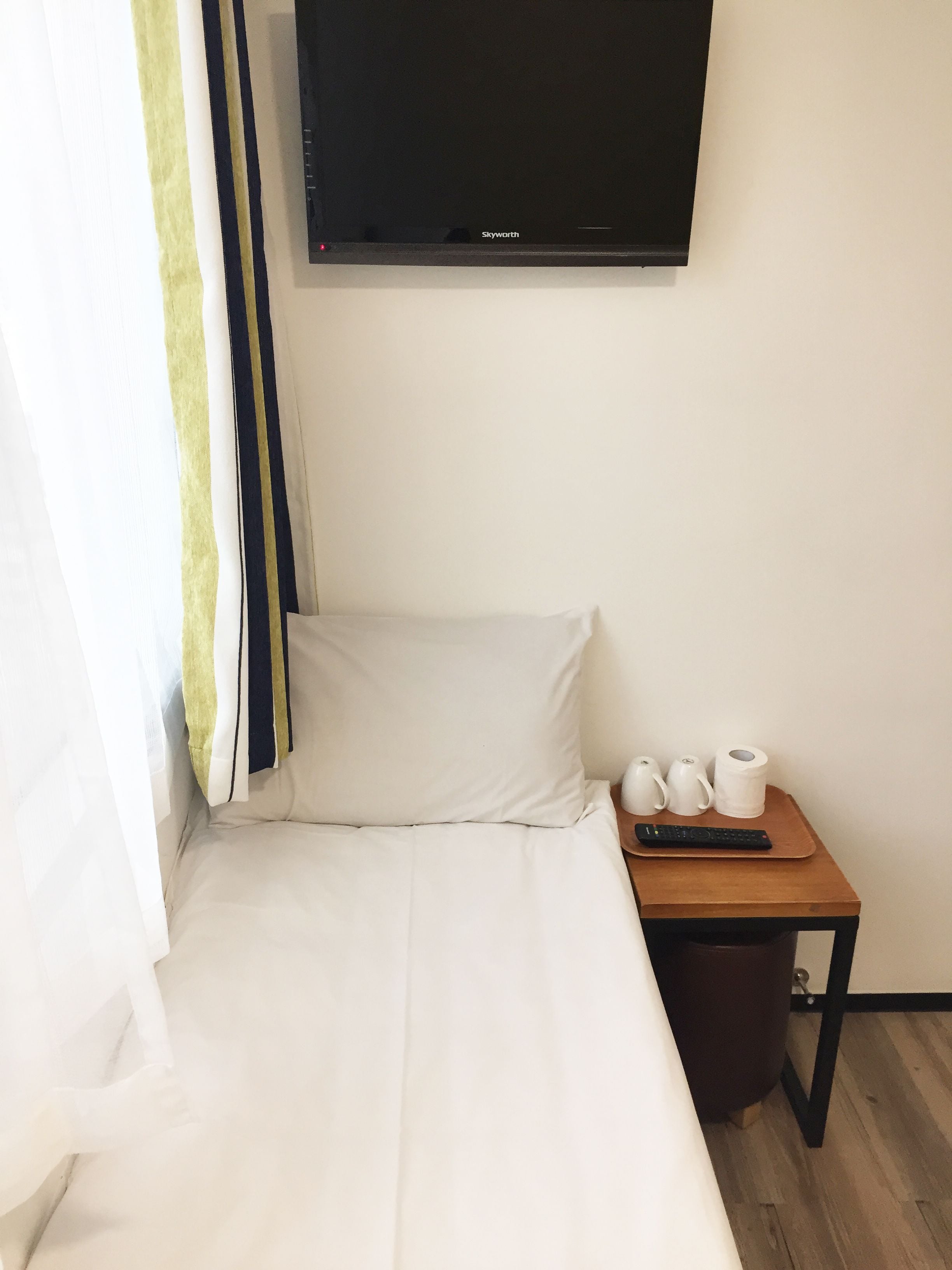 Une chambre d’hôtel avec un lit, un téléviseur fixé au mur, une petite table avec une télécommande et un rideau.