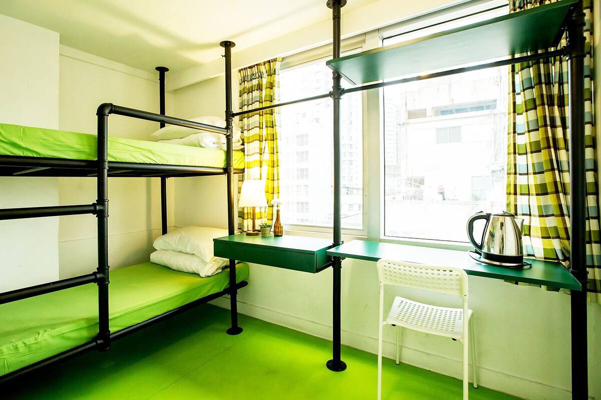 A bunk bed room with a desk and chair, a window with curtains, and a kettle on the desk.