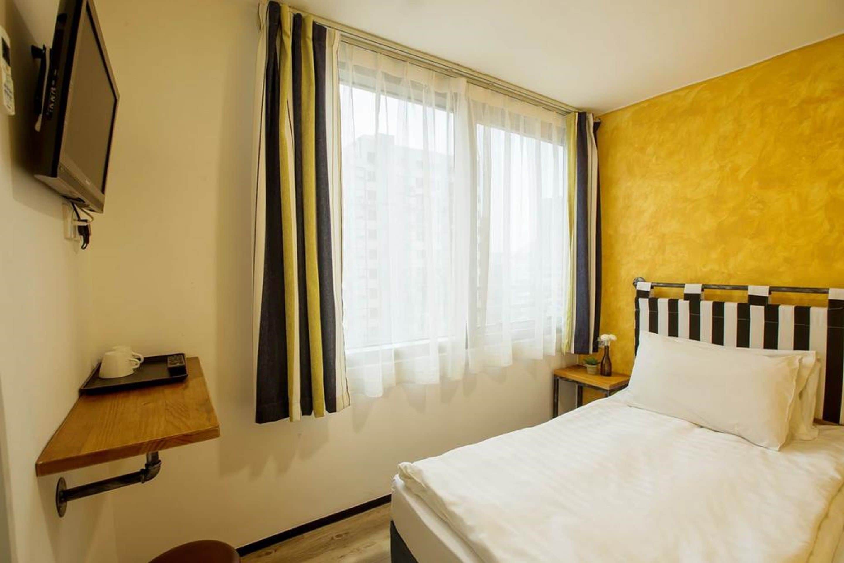 A hotel room with a bed, a television on the wall, a window with curtains, a small table with a cup, and a nightstand with a lamp.