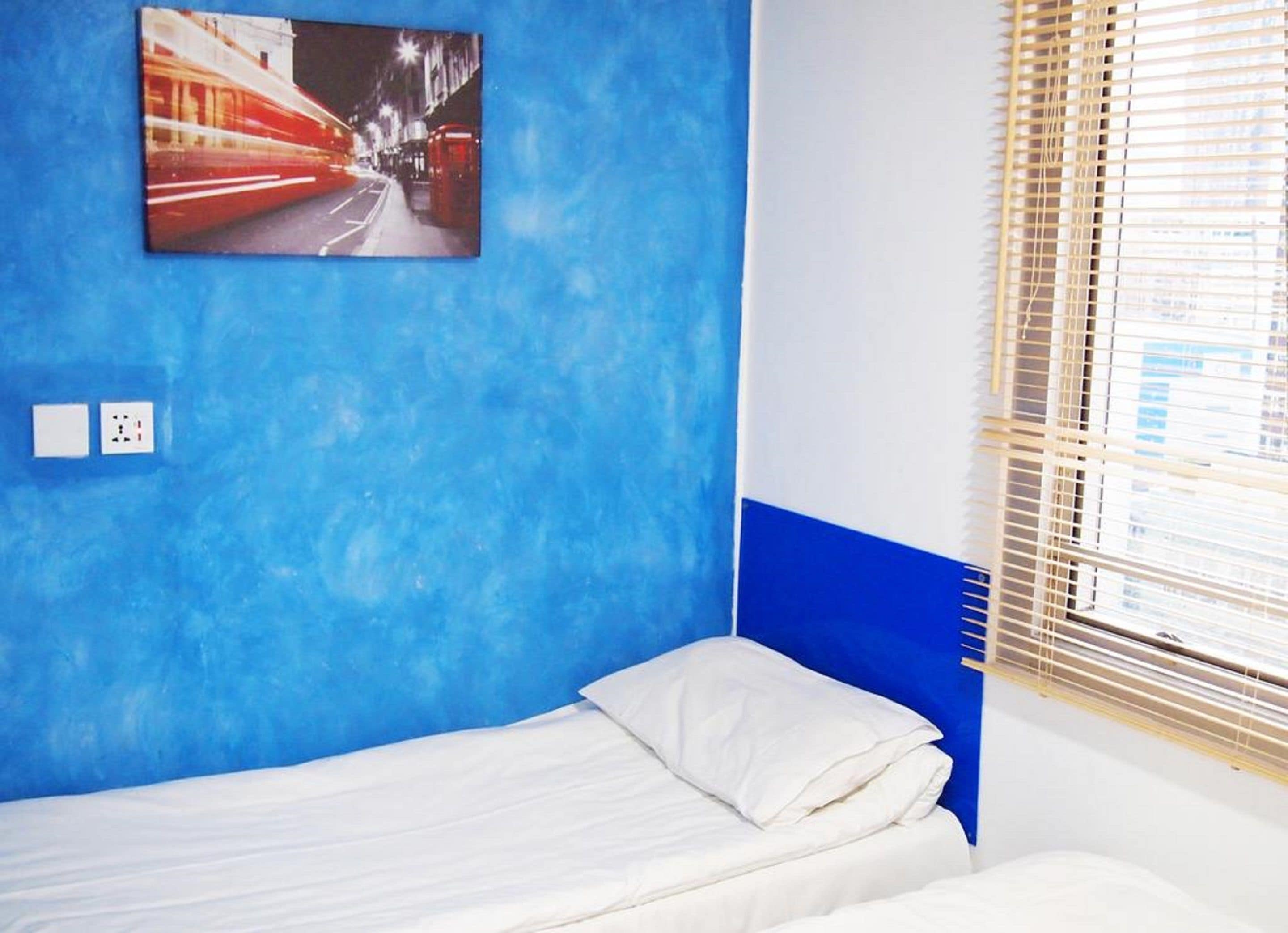 A room with a bed, a blue accent wall, a window with blinds, and a picture of a city street.