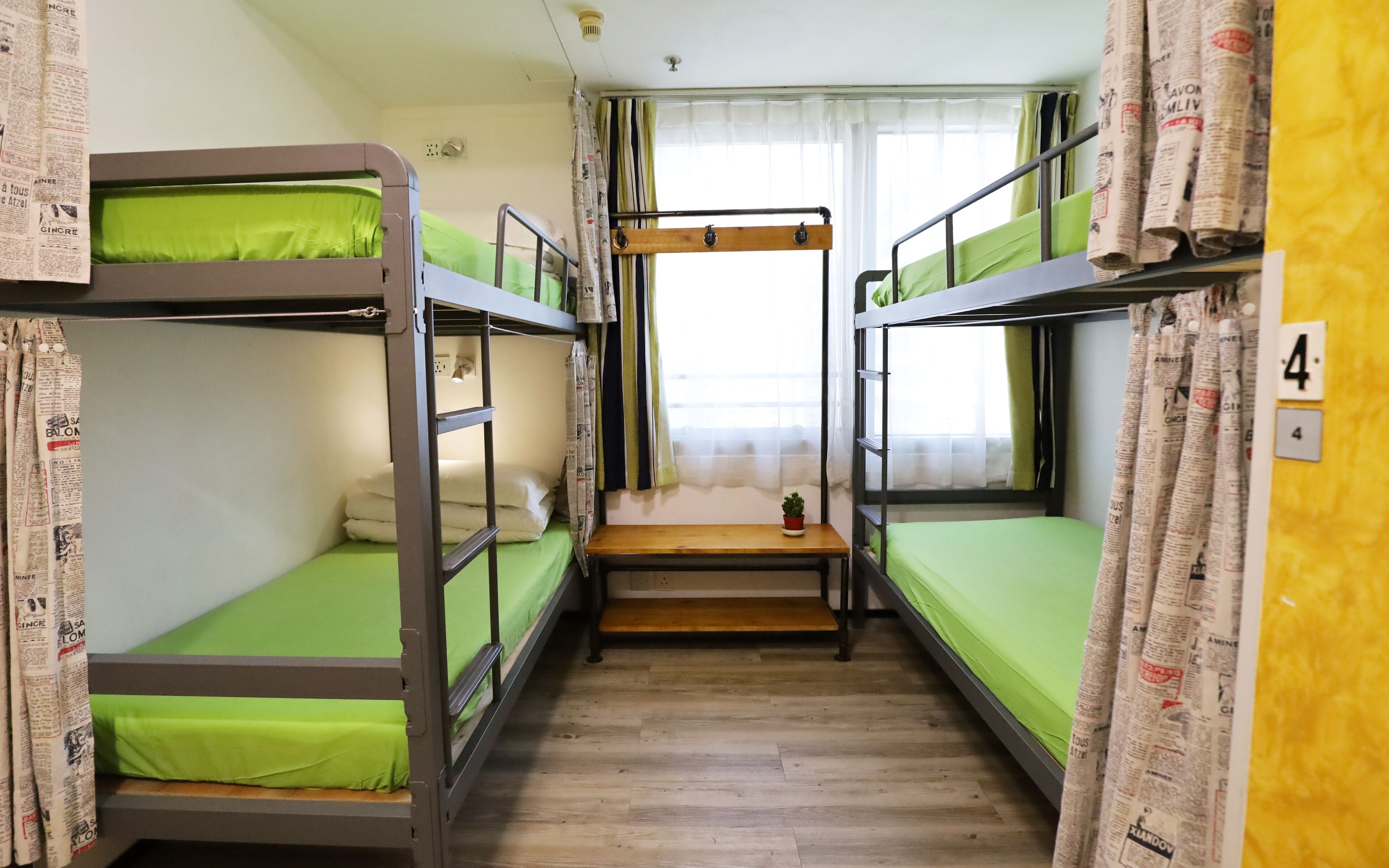 A dormitory room with bunk beds, a small wooden table, and a window with curtains.