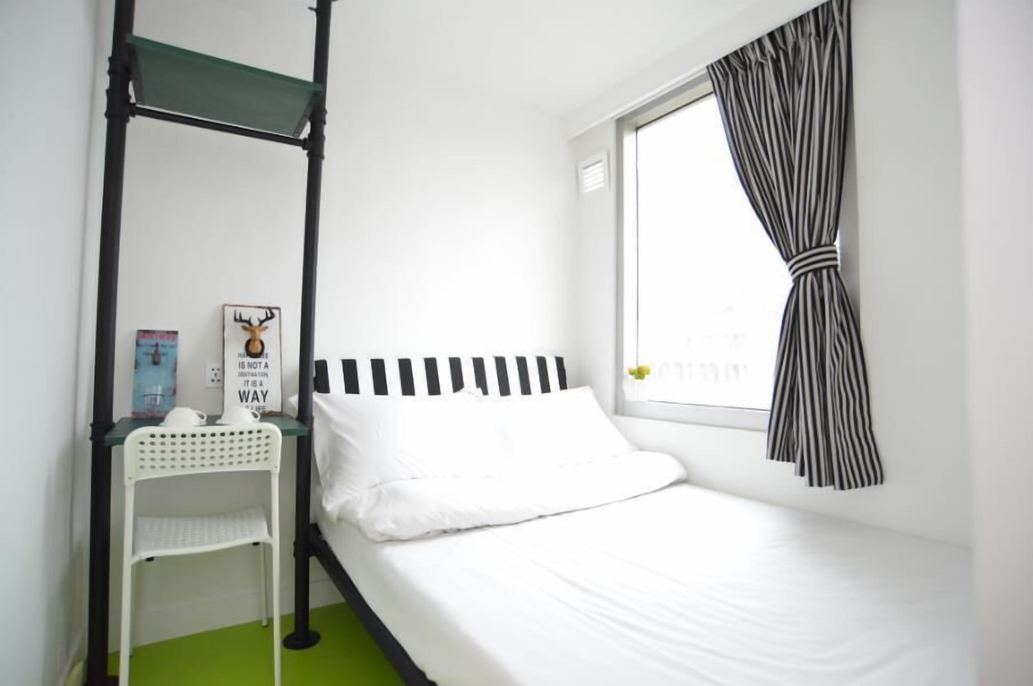 A single bed with a black and white striped headboard, a small bedside table with a white chair, a window with black and white striped curtains, and a wall-mounted shelf with a framed picture and a small plant.