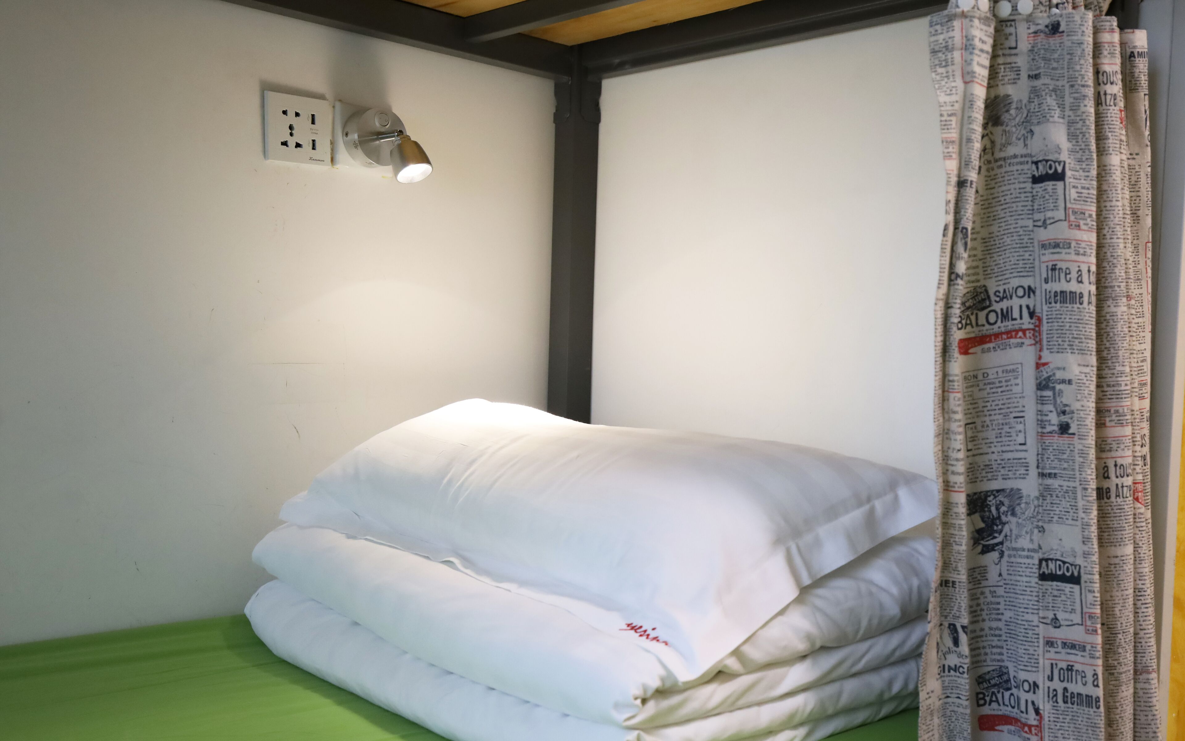 Ein kleines Zimmer mit einem Einzelbett, einer grünen Matratze, einem weißen Kopfkissen, einer Wandleuchte und einem Vorhang mit Zeitungsdruck.