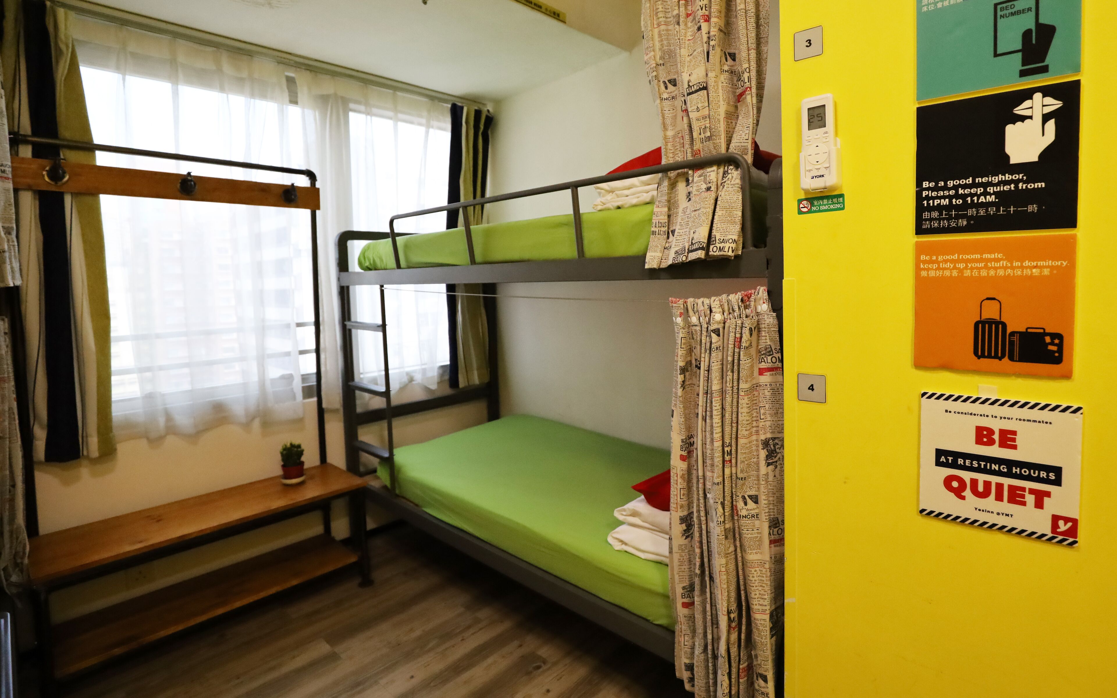 Une chambre de dortoir avec des lits superposés, une petite table et une fenêtre avec des rideaux légers. On y trouve des affiches murales avec des messages tels que « RESPECTEZ LE CALME PENDANT LES HEURES DE REPOS » et « Soyez un bon colocataire ».