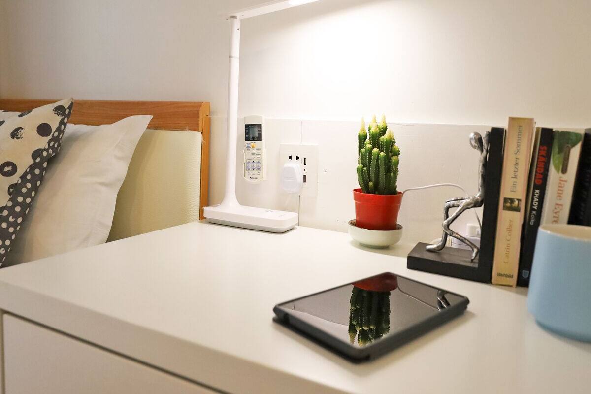 A neatly arranged hotel room with a bed, bedside table, a red potted plant, a bedside lamp, a digital scale, and a tablet.