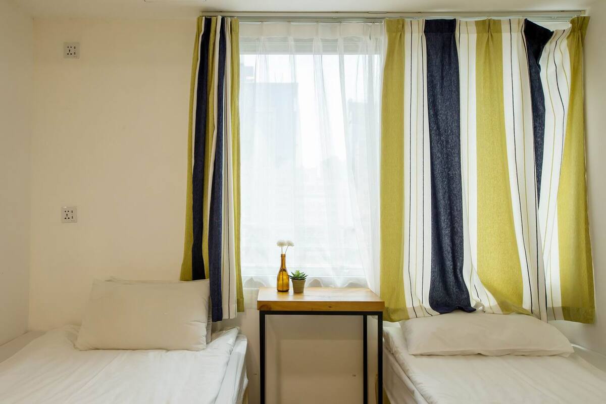 A hotel room with two single beds, a small table with a vase and a plant, and a window with striped curtains.