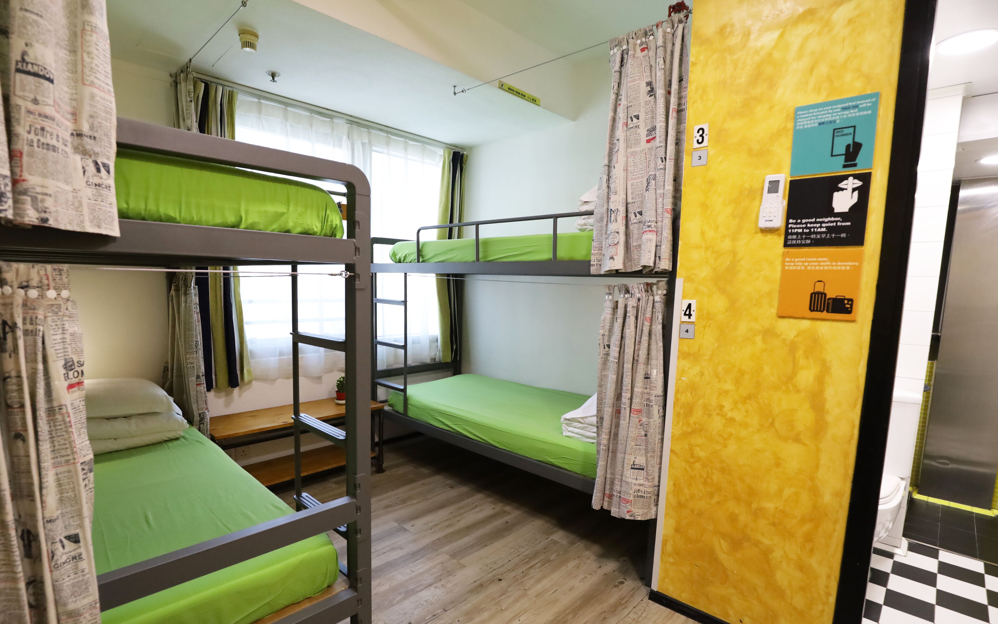 A dormitory room with bunk beds, a small desk, and a window with curtains.