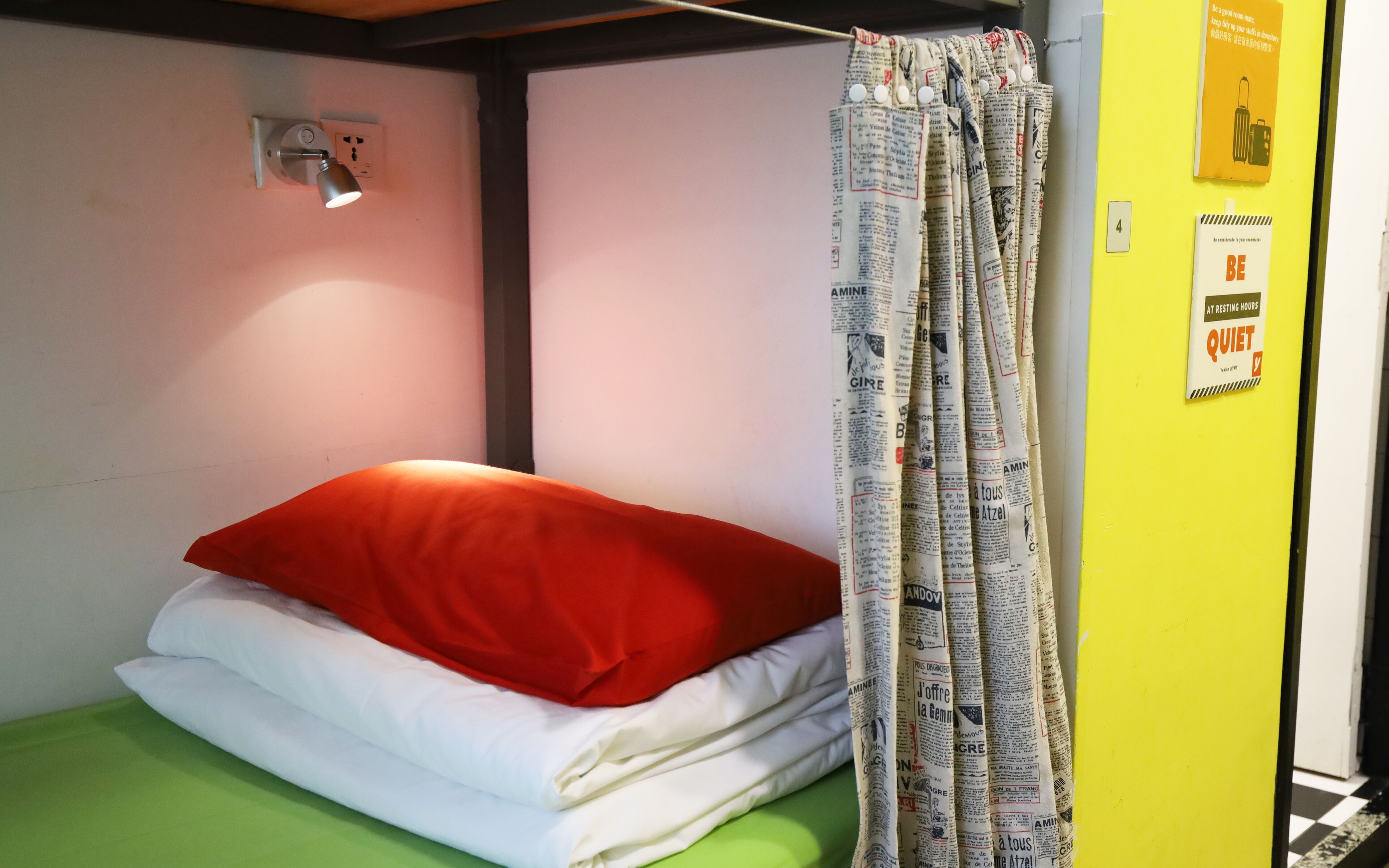 Uma cama de solteiro com travesseiro vermelho e lençóis brancos, uma cortina feita de jornal, uma luminária na parede e um aviso com a inscrição 'BE QUIET'.