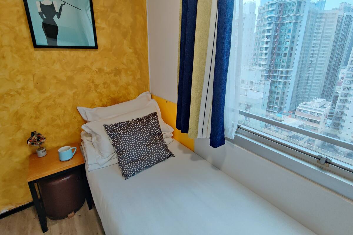 A hotel room with a bed, a nightstand, a framed picture, and a window with a city view.