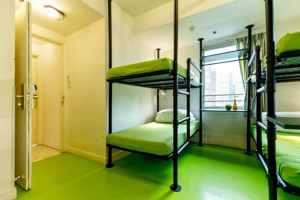 A dormitory room with bunk beds, a window with a view of buildings, and a door with a handle.