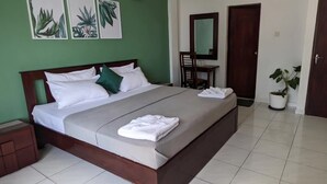 A neatly made bed with white linens and a grey bedspread, a wooden nightstand with a drawer, a chair with a mirror, and a potted plant. - Palmyrah Residencies (Colombo)
