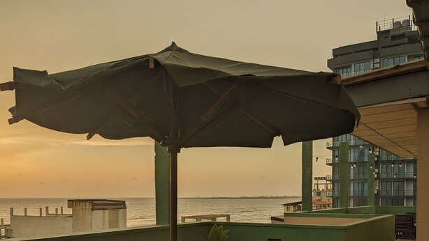 A wooden table and chairs under a large umbrella on a balcony with a view of the sea and a building in the background. - Palmyrah Residencies (Colombo)