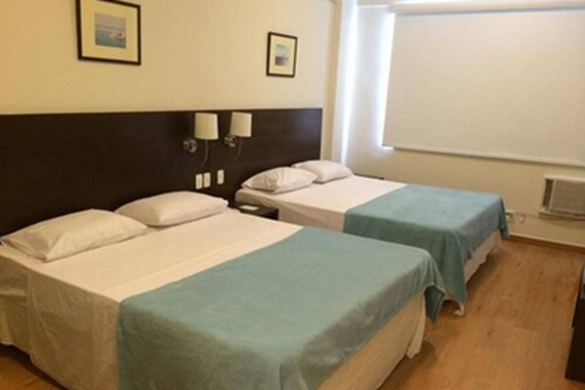 Une chambre d’hôtel avec deux lits, chacun doté d’une tête de lit blanche et d’une couvre-lit bleue. Deux lampes murales sont fixées sur la tête de lit, et il y a une fenêtre avec des stores.