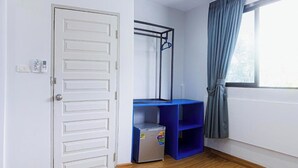 Una habitación con una puerta blanca, un escritorio azul con estantes, un refrigerador pequeño y una ventana con cortinas.