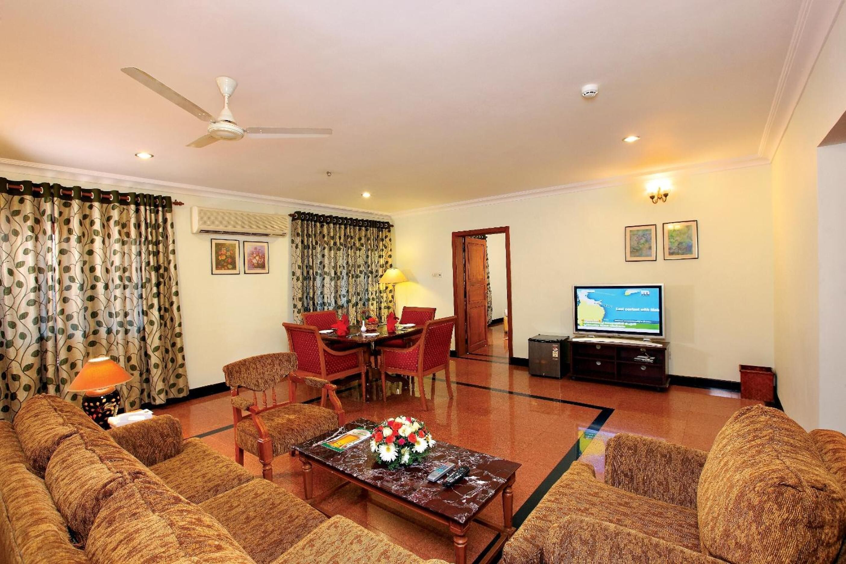 A living room with a sofa, a coffee table with a floral centerpiece, a dining table with chairs, a television, and a ceiling fan.