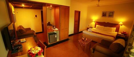 A hotel room with a bed, a sofa, a dining table with flowers, a kitchen area, and a ceiling fan.