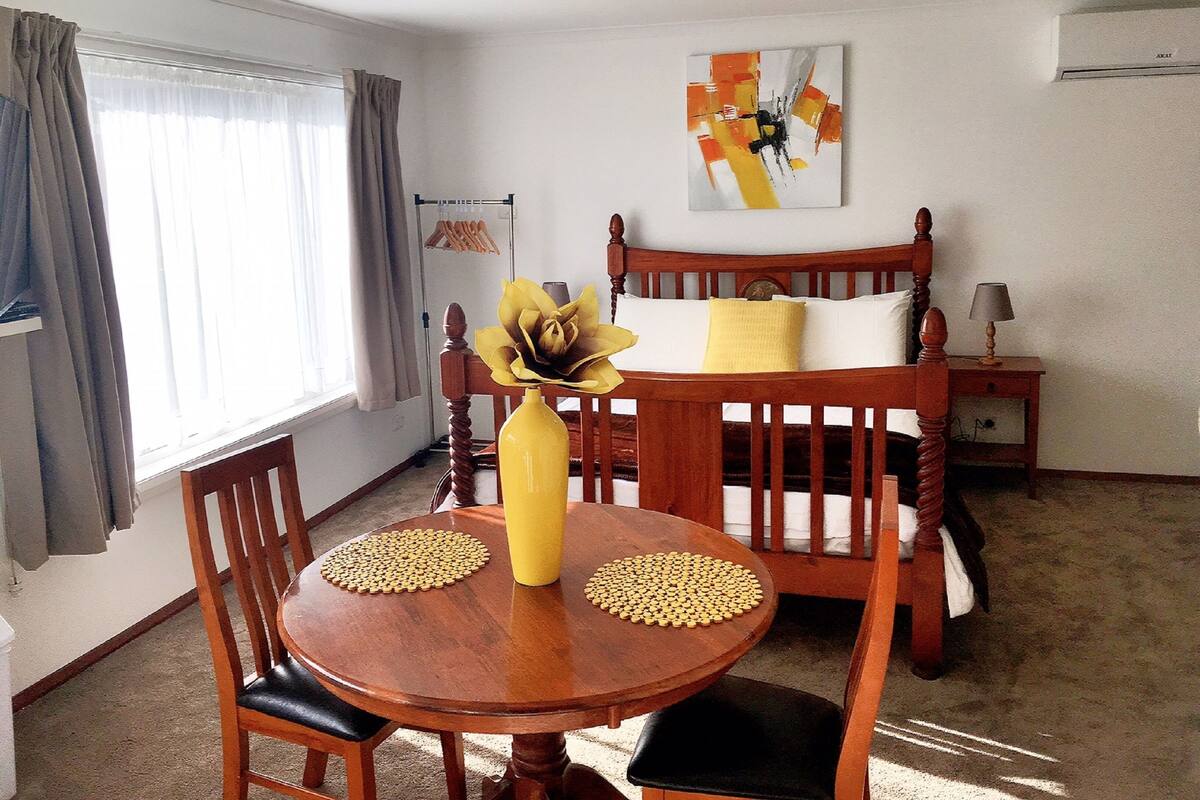 A room with a wooden bed, a round wooden table with a yellow vase, and a painting on the wall.