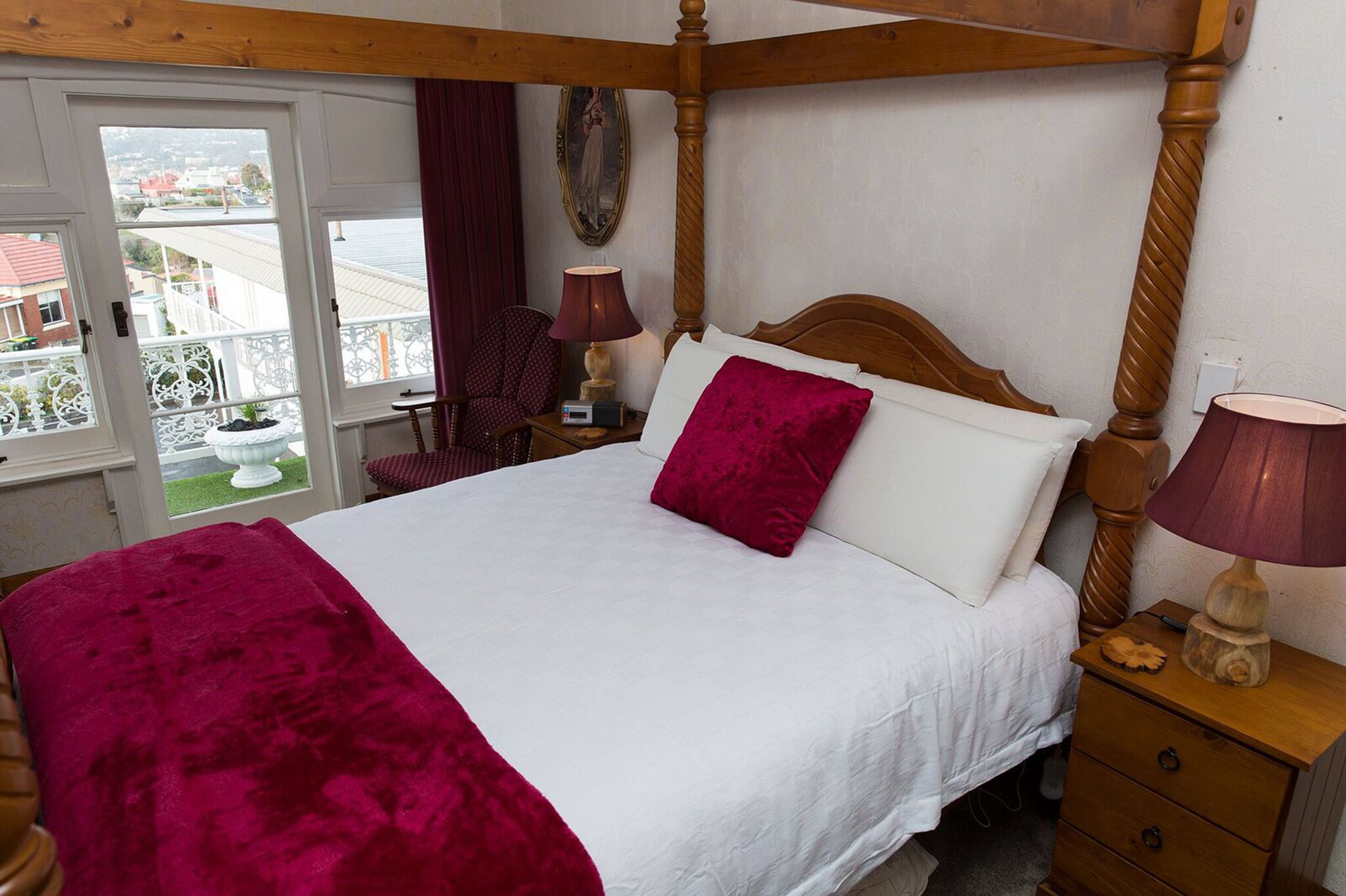 Un lit à baldaquin avec une couvre-lit blanche et un plaid rouge. On aperçoit une table de chevet en bois avec une lampe, ainsi qu’une petite table sur laquelle est posé un objet décoratif. Une chaise se trouve dans un coin, et une fenêtre donne sur des bâtiments.