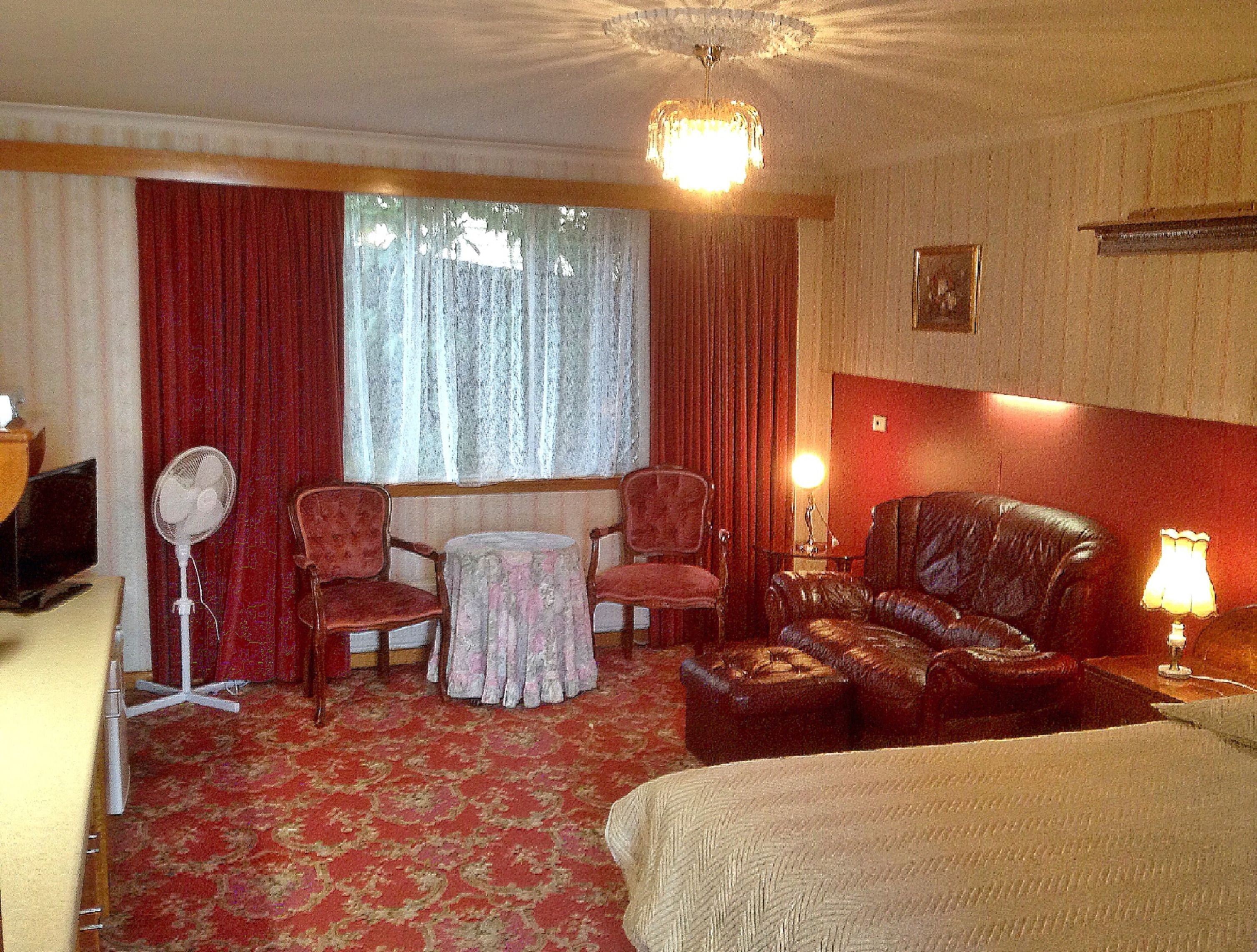 Habitación de hotel con cama, sofá de cuero, dos sillones, una mesita, un ventilador y una ventana con cortinas.