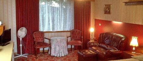 Habitación de hotel con cama, sofá de cuero, dos sillones, una mesita, un ventilador y una ventana con cortinas.