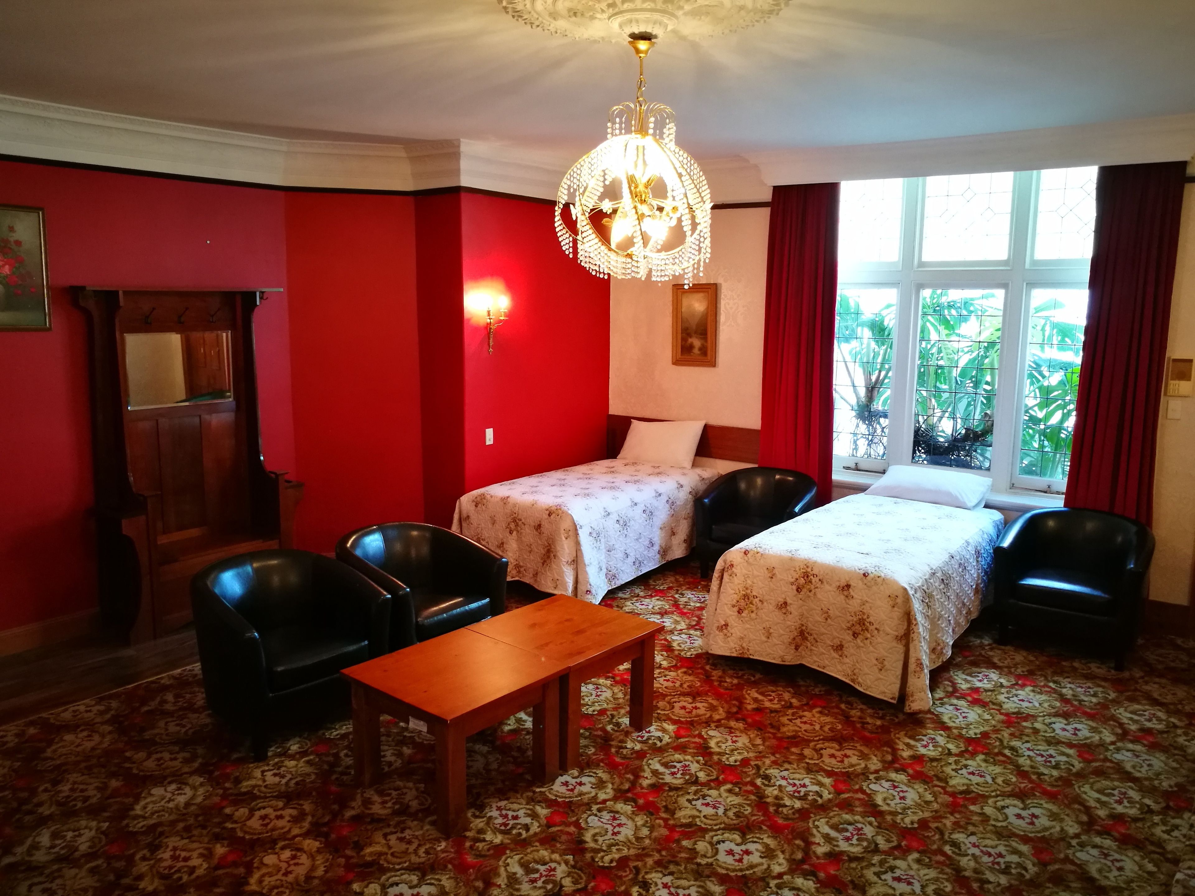 Una habitación con dos camas, una mesa de madera, sillas negras, una araña y una ventana con cortinas rojas.