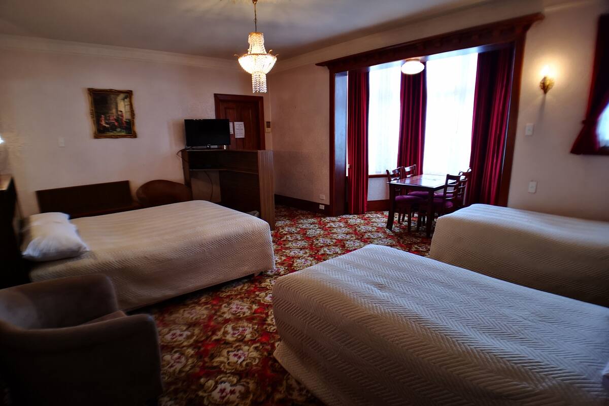 A hotel room with two single beds, a desk with a TV, a dining table with chairs, and a chandelier.