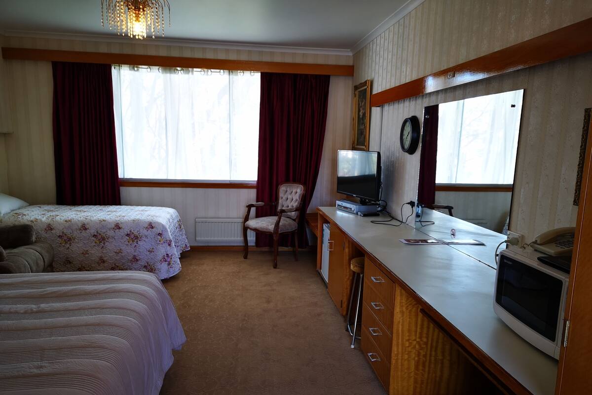 A hotel room with two beds, a desk with a television, a chair, and a large window with curtains.