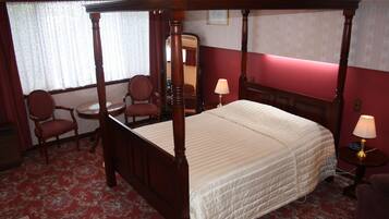 A four-poster bed with a quilt, a round table with a chair, and a mirror on the wall.