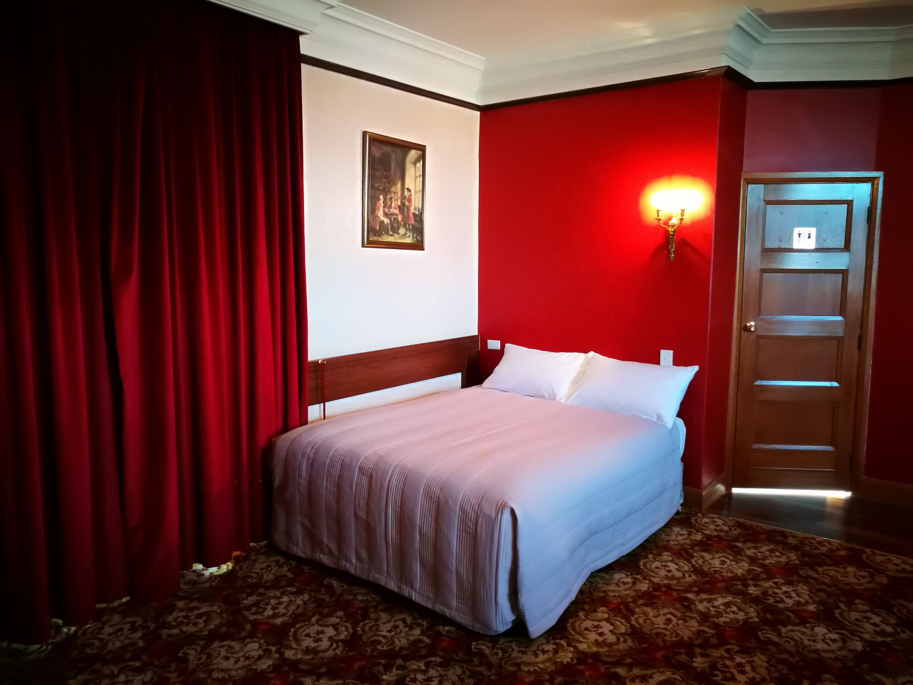 Habitación de hotel con cama, cortinas rojas, una lámpara fijada a la pared, una puerta y alfombra estampada.