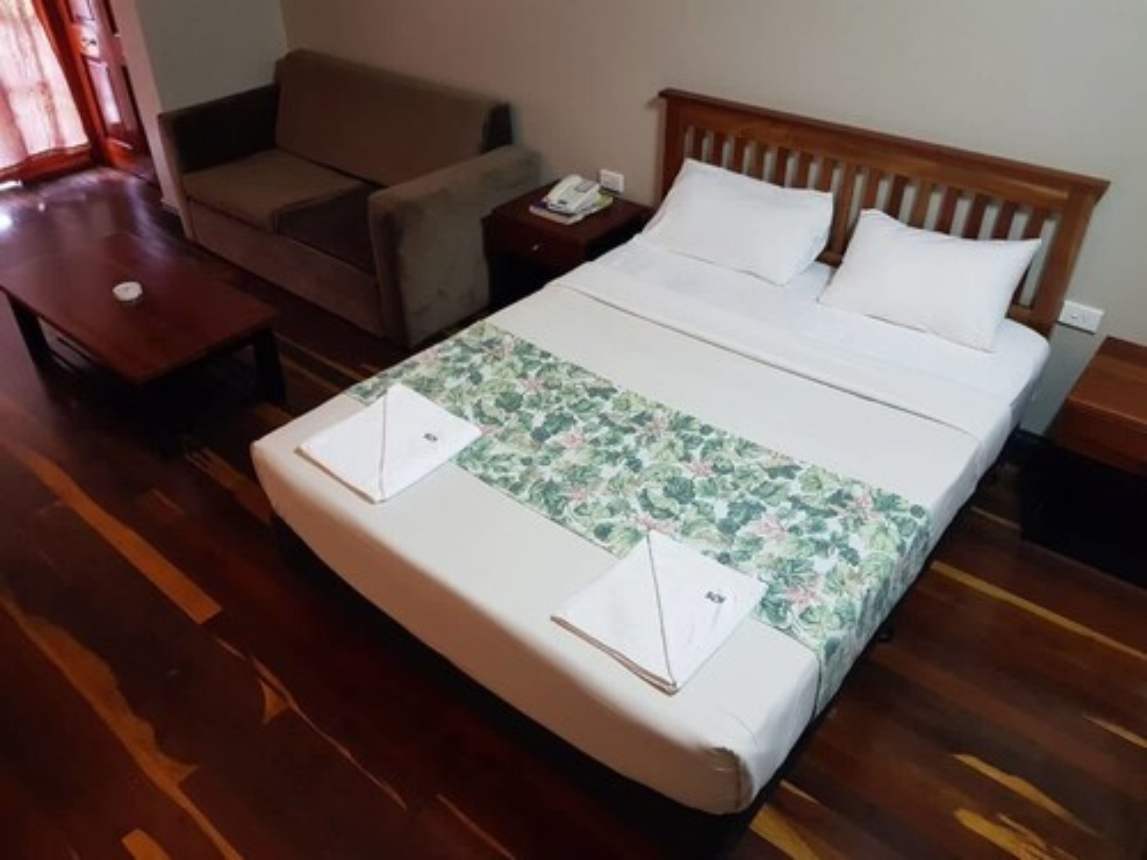 A hotel room with a wooden floor, a bed with white bedding, a floral-patterned bedspread, a wooden headboard, a nightstand, a chair, and a small table with a book and a remote control.