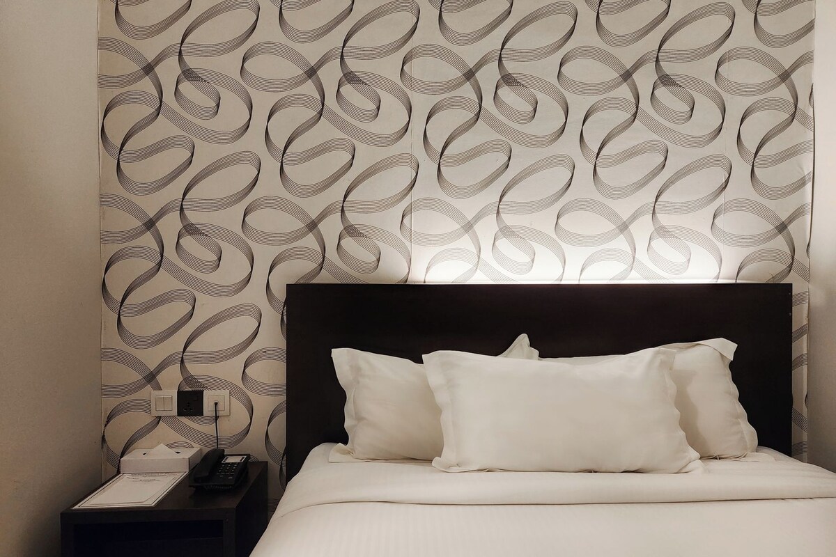 Une chambre d’hôtel avec un papier peint à motifs, une tête de lit noire, du linge de lit blanc et une table de chevet avec un téléphone et une lampe.