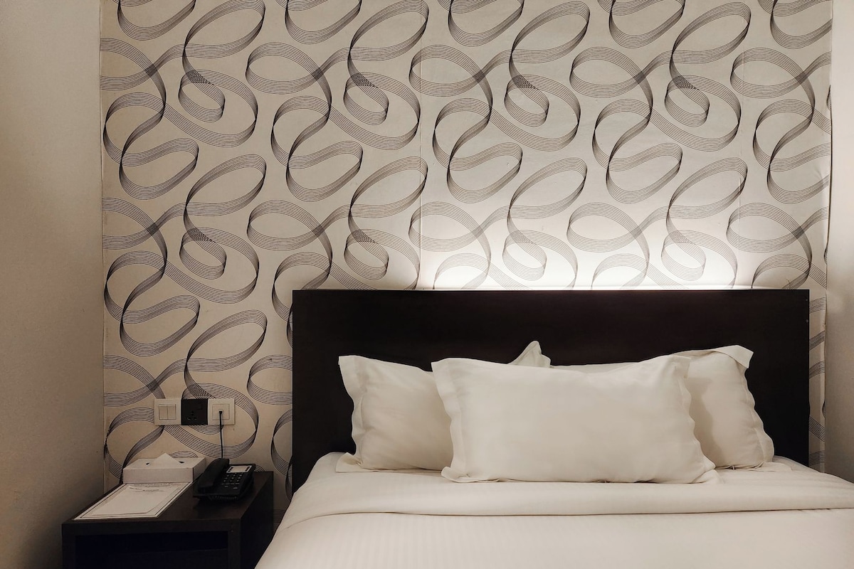 Ein Hotelzimmer mit gemustertem Wandteppich, einem schwarzen Kopfteil, weißer Bettwäsche und einem Nachttisch mit Telefon und Lampe.