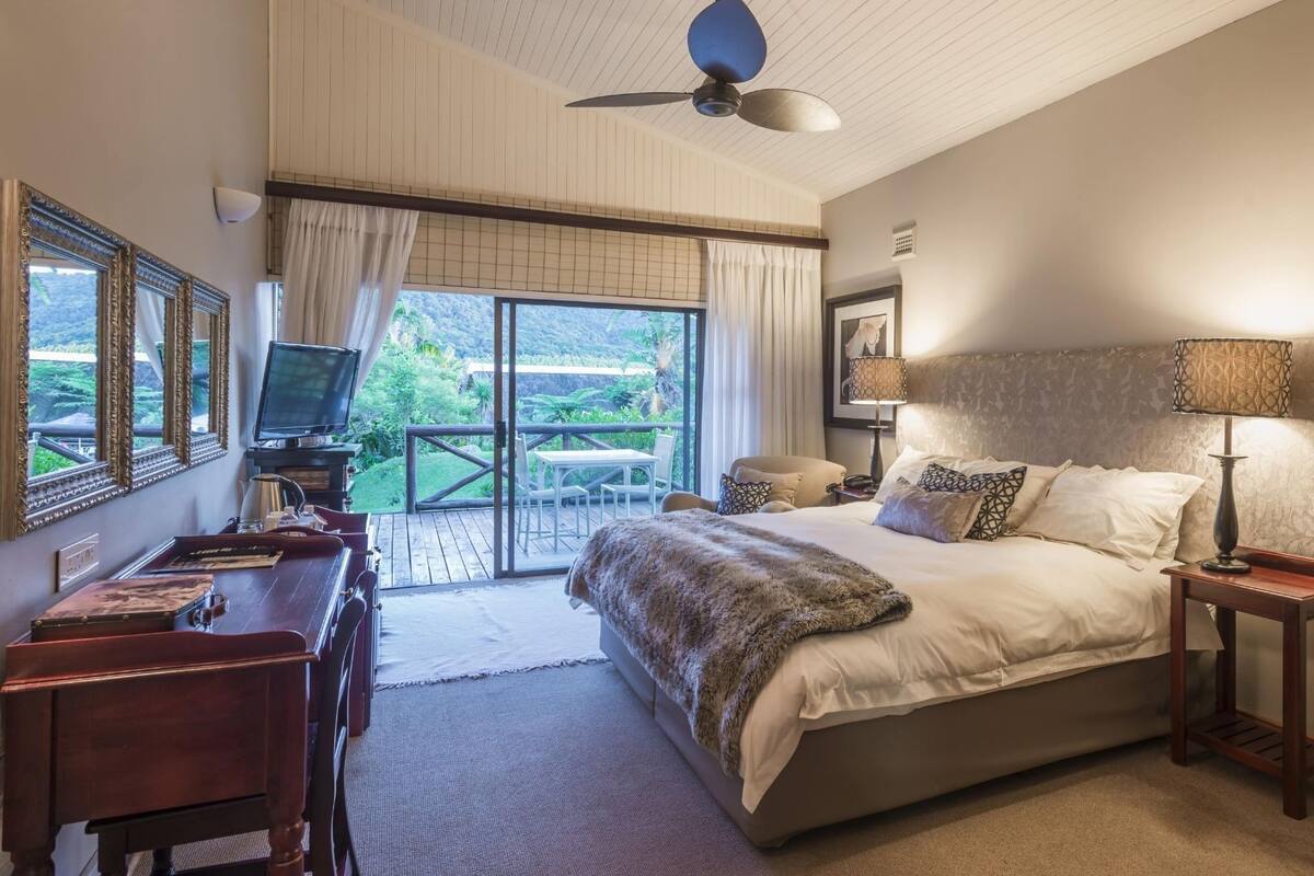 Un dormitorio con una cama grande, un escritorio, un televisor y un balcón con vistas a zonas verdes.