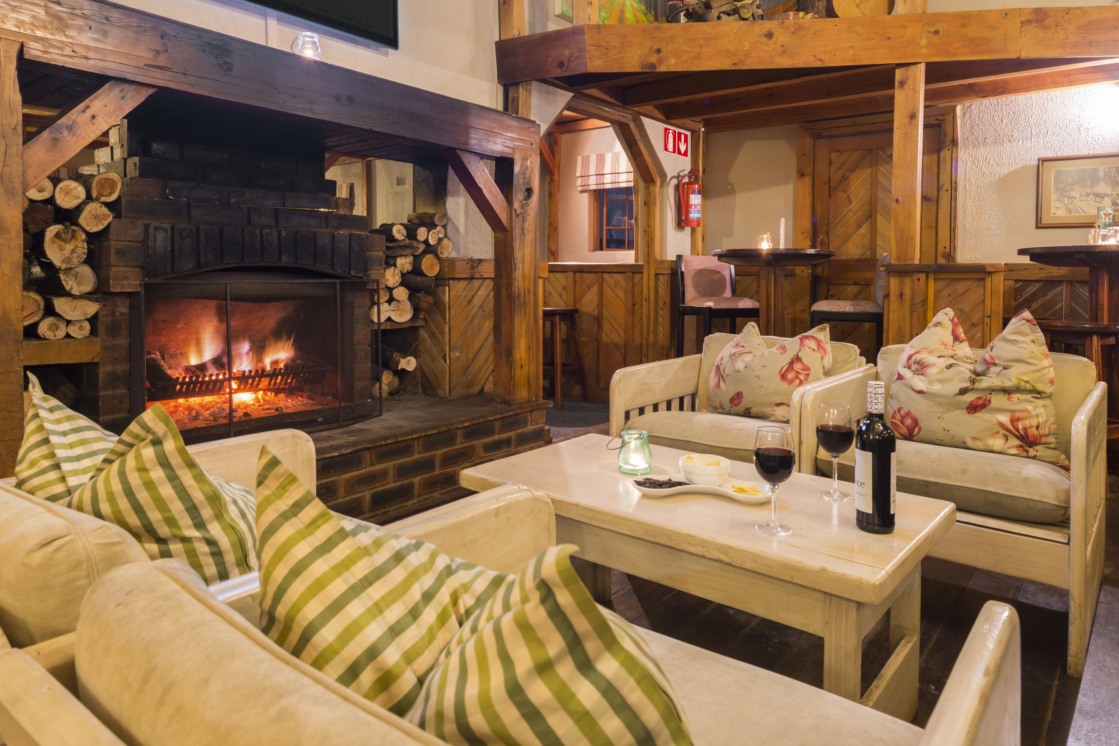 Una habitación acogedora con chimenea, vigas de madera, un sofá con cojines estampados y una mesa con copas de vino y una botella.