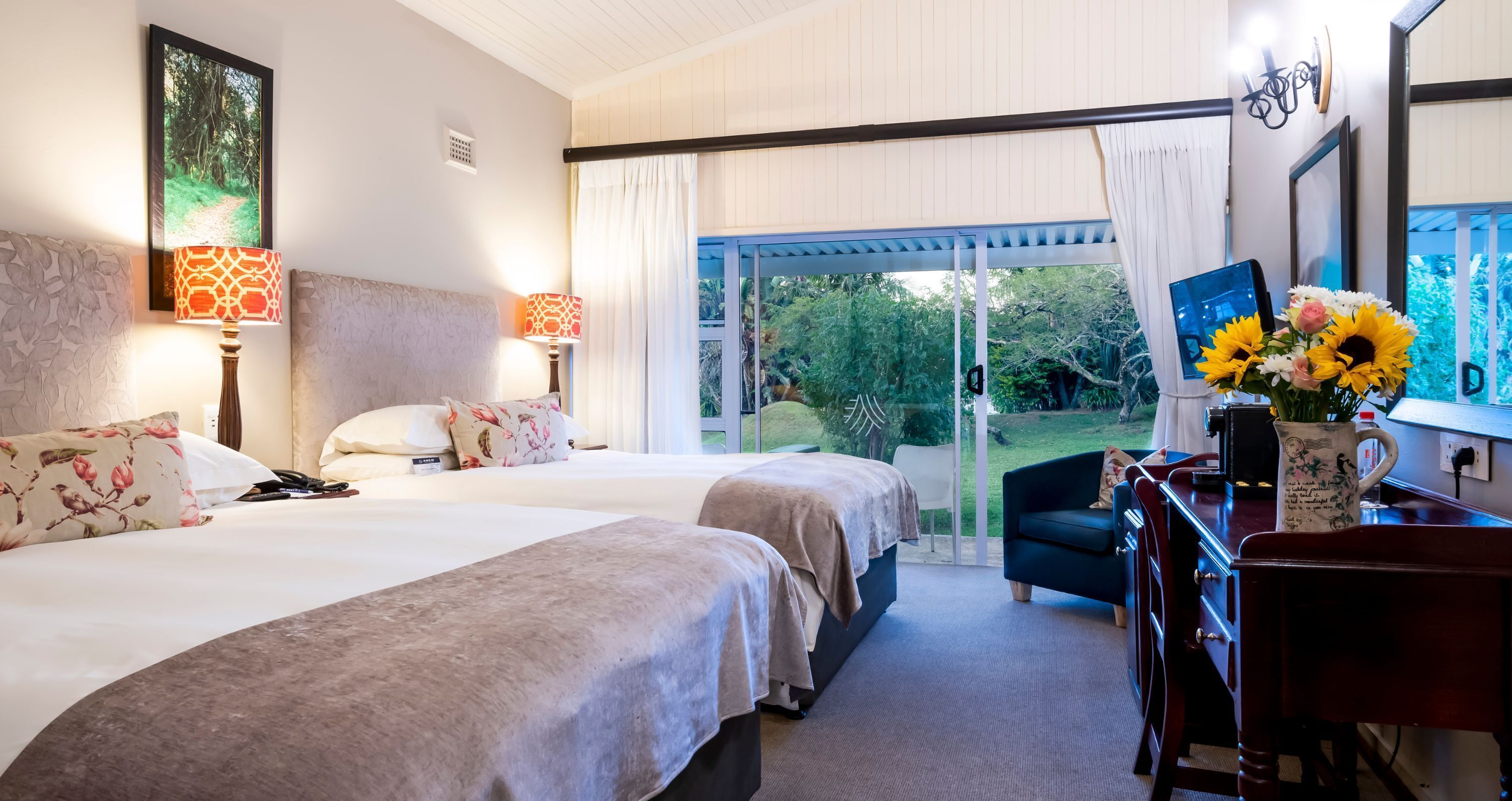 A hotel room with two beds, a desk, a chair, a TV, and a view of the outdoors.