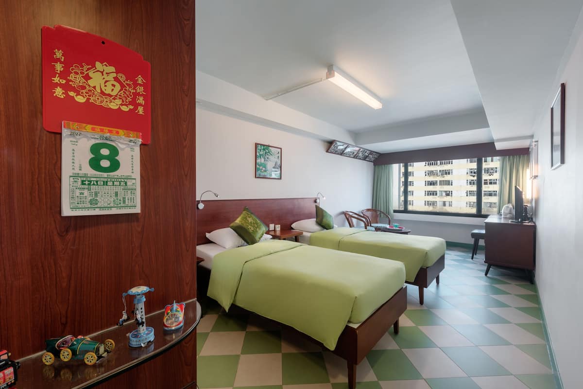 A hotel room with two beds, a wooden headboard, a small desk, a chair, a window with a view of buildings, and a shelf with toys.