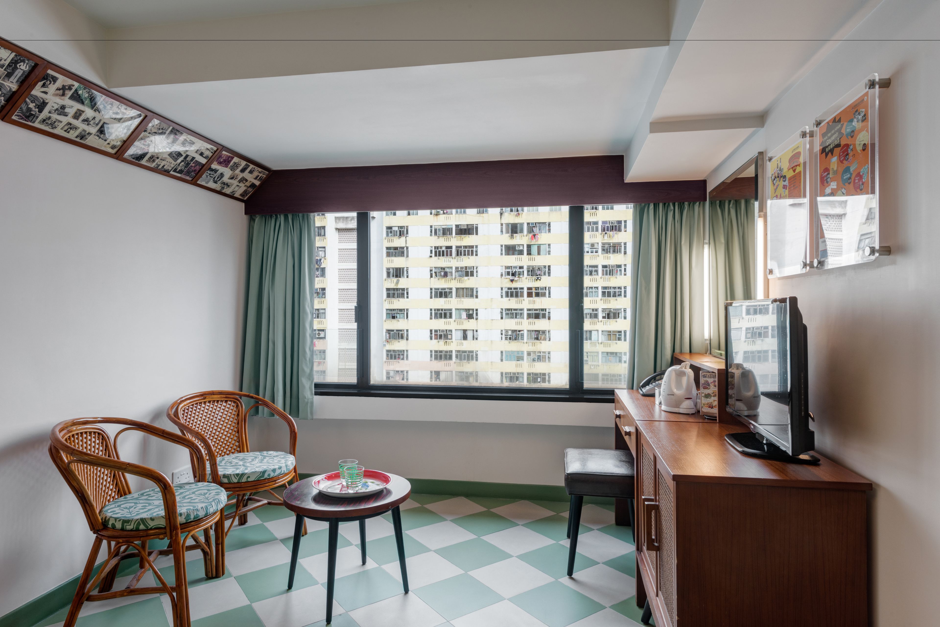 A room with a checkered floor, two wicker chairs, a small table with a glass and a tray, a wooden cabinet with a television, and a window with a view of a building.