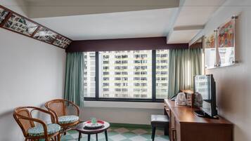 A room with a checkered floor, two wicker chairs, a small table with a glass and a tray, a wooden cabinet with a television, and a window with a view of a building.