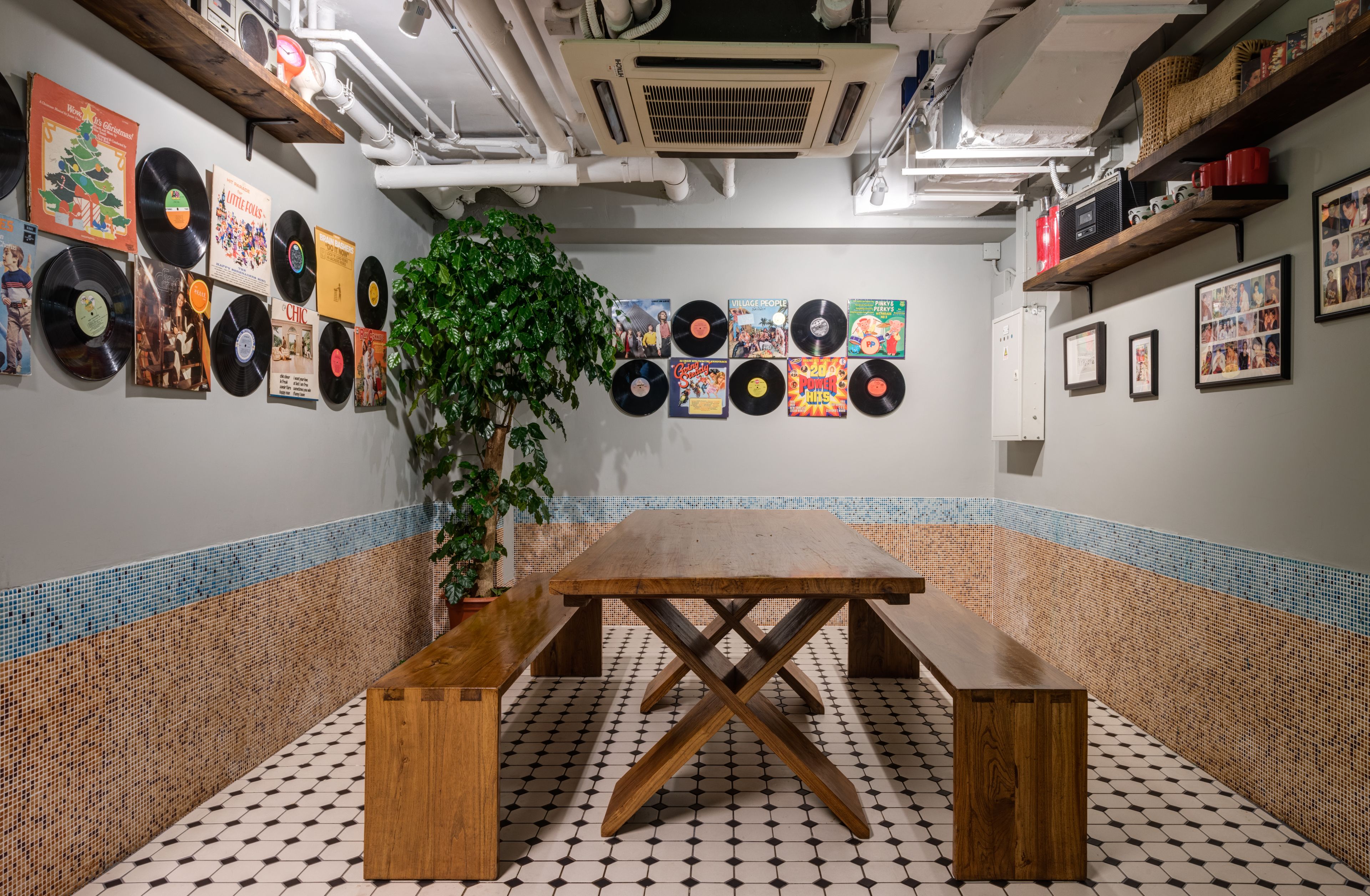 Una habitación con una mesa de madera y bancos, una pared decorada con discos de vinilo y fotografías enmarcadas, y una planta en maceta.